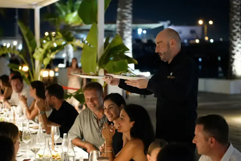 A waiter serves guests at a long outdoor dinner table under string lights at Rigatoni Ibiza.
