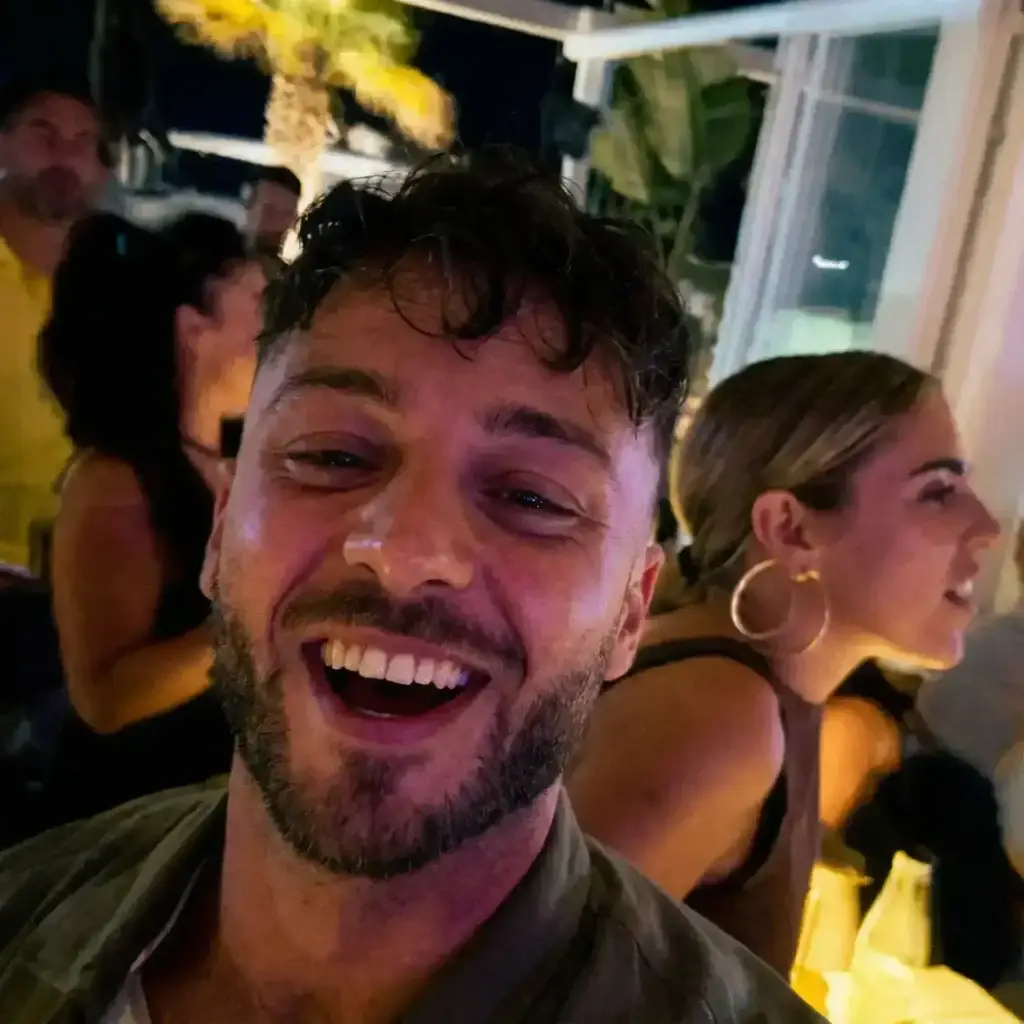A smiling man takes a selfie at a lively Ibiza nighttime gathering with people socializing behind.