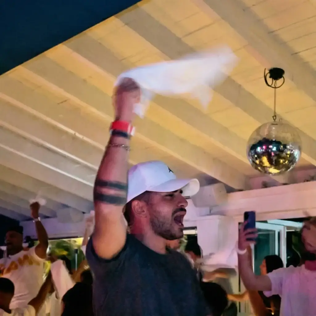 Man in white cap cheering at Rigatoni Ibiza party, people waving napkins, disco ball overhead.