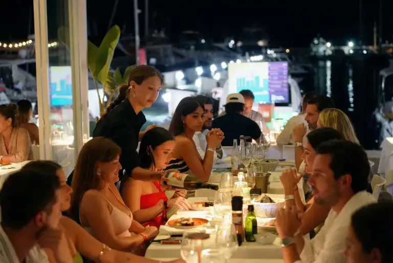A waitress serves food to a group dining at a lively waterfront Rigatoni Ibiza restaurant at night.