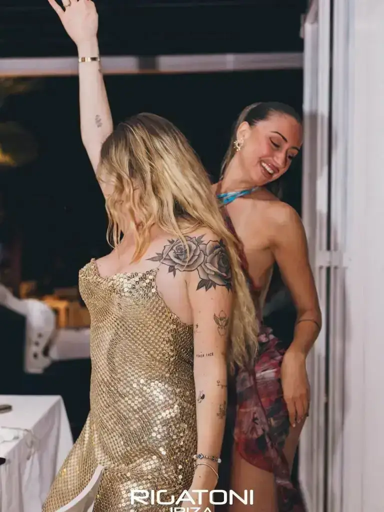 Two women dancing and smiling at a lively Rigatoni Ibiza event, one in gold, the other in a floral skirt.
