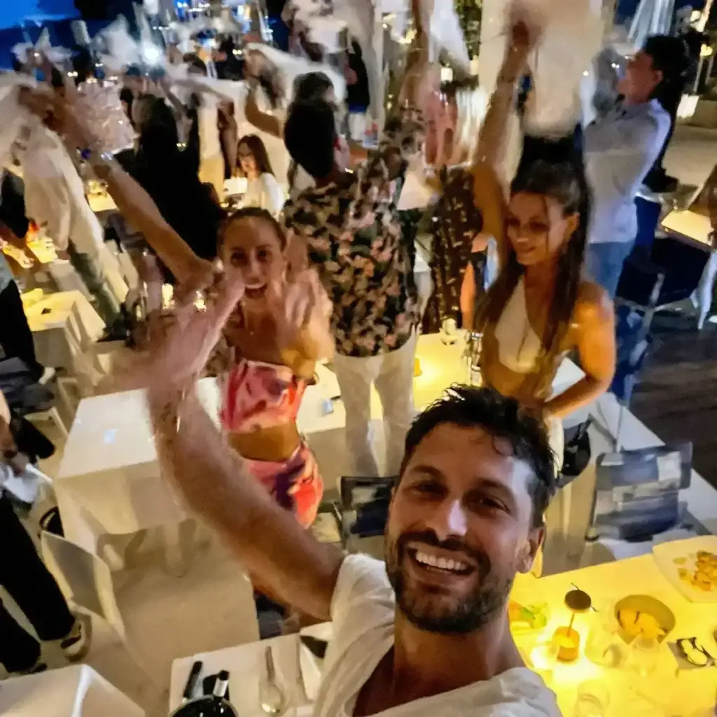 A group of people smiling and celebrating at a lively Ibiza restaurant, waving napkins at Rigatoni.