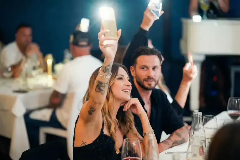 A woman and man at Rigatoni Ibiza raise phones with flashlights on, surrounded by other diners.