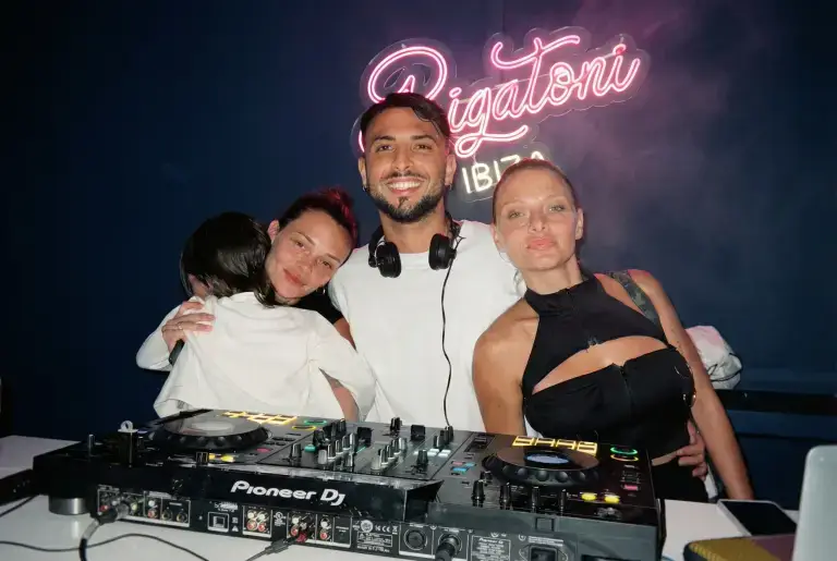 Three people smile behind DJ equipment with a glowing Rigatoni neon sign lighting up the Ibiza night.