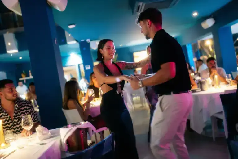 A couple dances joyfully in a lively blue-lit Ibiza restaurant while diners enjoy their meals at Rigatoni.
