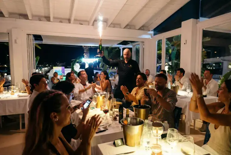 A waiter holds a sparkling bottle as people celebrate at Rigatoni Ibiza with white decor.