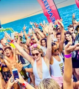 People celebrating at a lively Ibiza boat party, raising their arms with the ocean and banners behind.
