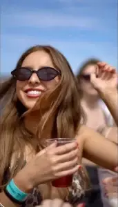 Woman in sunglasses smiling, holding a drink, and dancing at a sunny Ibiza boat party.