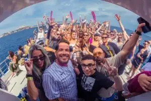 A large group enjoys a boat party in Ibiza, smiling and celebrating with ocean and coastline behind.