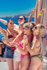 Five women in swimsuits smile and pose on an Ibiza boat party with blue sea and coastline behind.