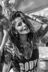 Woman with wavy hair and tattoos poses outdoors at an Ibiza boat party, sunlight on her face.