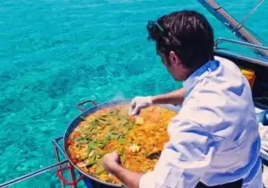 A chef in white prepares a large pan of paella on a boat party above Ibiza clear turquoise water.