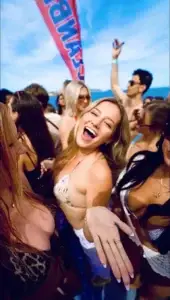 A smiling woman in a bikini shows off a ring at a lively Ibiza boat party with friends.