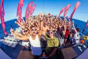 A large group dances and cheers at an Ibiza boat party with red flags and blue ocean behind.