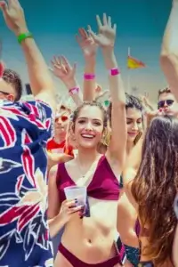 Young woman in a maroon bikini smiles and raises her arms at a lively Ibiza boat party.